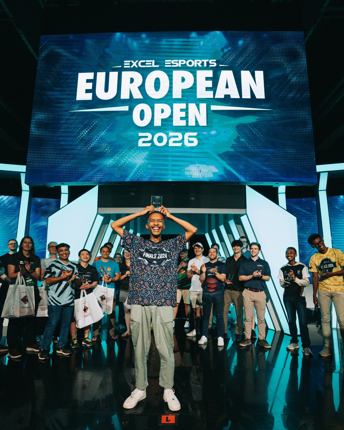 A student lifts a trophy overhead on stage, applauded by the rest of the field, beneath a screen reading Excel Esports European Open 2026.
